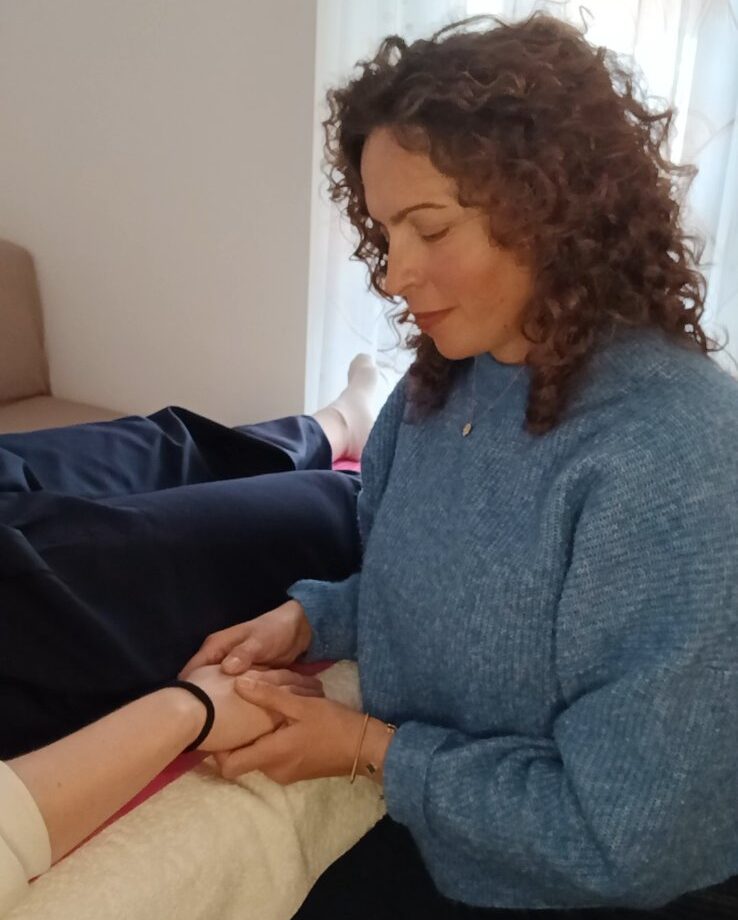 Η Ιουλία κρατά με τα δύο χέρια το πόδι θεραπευόμενου, σε ένα ήσυχο, οικείο μέρος της συνεδρίας.